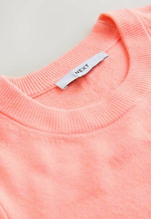Korallrosa tröja med ribbad halsringning, mjuk textur och en etikett med texten "NEXT" fäst på insidan av halsringningen.