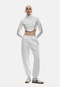 Witte cropped zip-up jack met zwarte zijstrepen, gecombineerd met bijpassende witte joggerpants met elastische boorden, gedragen met sneakers.