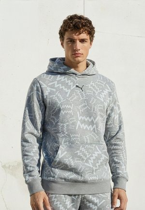 Jeune homme portant un sweat à capuche gris et un pantalon de jogging assorti avec des motifs en zigzag blancs, debout devant un mur blanc uni.