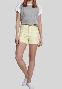 Unos pantalones cortos de mezclilla amarillos con los puños enrollados, de cintura alta y con bolsillos frontales. Combinados con una blusa de manga corta en gris y blanco y zapatos blancos tipo slip-on.