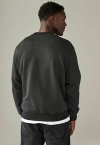 Sudadera gris oscuro con cuello redondo y puños acanalados, con una textura suave y lisa. Superpuesta sobre una camiseta blanca y combinada con jeans oscuros.