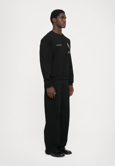 Felpa nera con collo rotondo, design ricamato, vestibilità rilassata, abbinata a pantaloni neri larghi e scarpe nere, su sfondo bianco.