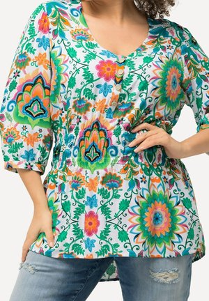 Vrouw draagt een kleurrijk bloemetjespatroon blouse met knopen, driekwart mouwen en versleten blauwe spijkerbroek, poseert met één hand op de heup.