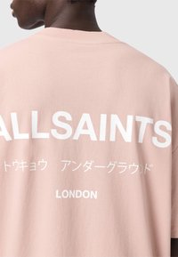 Φαρδιά ανοιχτό ροζ βαμβακερή μπλούζα. Φέρει μεγάλο λευκό κείμενο στην πλάτη: "ALLSAINTS," επιπλέον κείμενο σε άλλες γλώσσες και τη λέξη "LONDON."