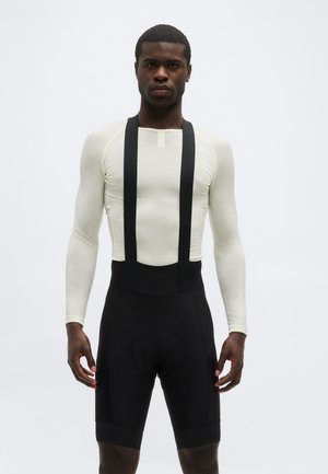 Uomo con indosso una maglia beige aderente a maniche lunghe e pantaloncini da ciclismo neri con bretelle, in piedi davanti a uno sfondo semplice.