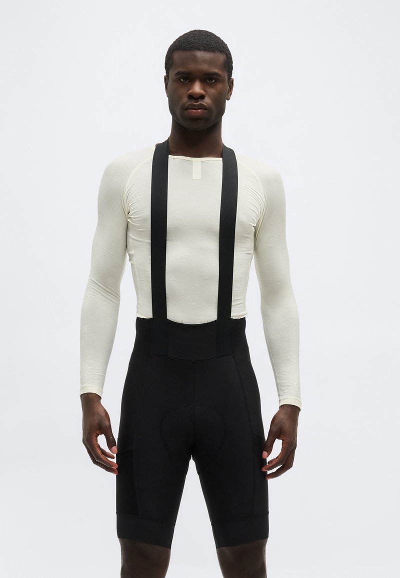 Άνδρας που φοράει εφαρμοστό μακρυμάνικο μπεζ μπλουζάκι και μαύρα σορτς ποδηλασίας με τιράντες, στέκεται μπροστά σε απλό φόντο.