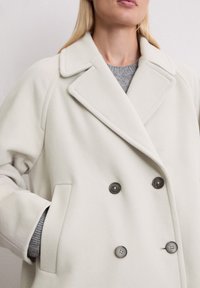 Manteau beige à double boutonnage fabriqué dans un matériau doux, avec des revers crantés, deux poches avant et des boutons foncés pour la fermeture.