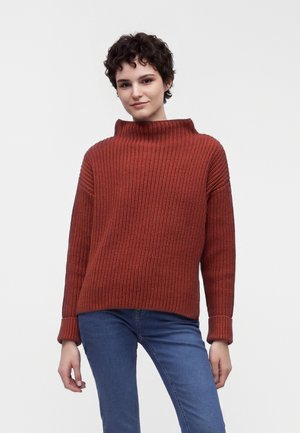 Jong persoon met kort krullend haar, gekleed in een rood gebreide trui en blauwe spijkerbroek, staand tegen een effen lichte achtergrond.