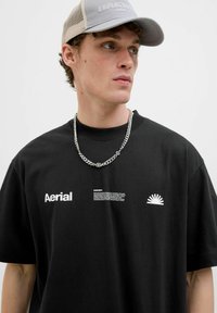 Camiseta de algodón negra de manga corta con el texto blanco "Aerial" y un gráfico de un amanecer en el lado izquierdo del pecho. Collar de cadena plateada visible.