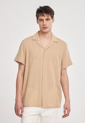Camicia beige a maniche corte con bottoni, realizzata in tessuto testurizzato, con colletto e cinque bottoni sulla parte anteriore. Indossata con pantaloni di colore chiaro.