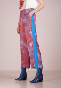 Pantalones de pierna ancha en rojo y azul con un patrón de hojas de palma, complementados por una franjas lateral azul y bolsillos laterales. La textura parece suave.