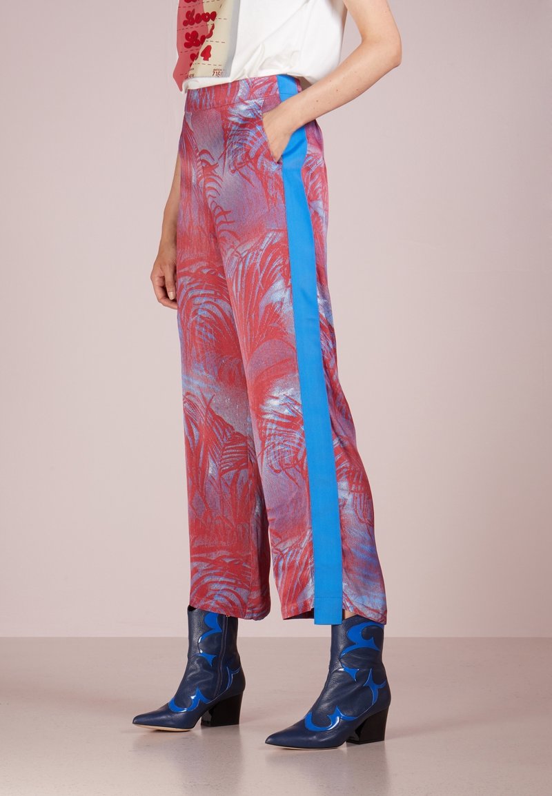 Pantalones de pierna ancha en rojo y azul con un patrón de hojas de palma, complementados por una franjas lateral azul y bolsillos laterales. La textura parece suave.