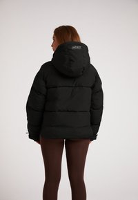 Zwarte pufferjack met een grote capuchon, voorzien van een gestikt ontwerp en elastische mouwuiteinden. Logo zichtbaar op de achterkant ter hoogte van de nek.