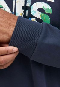 Marineblåt sweatshirt med ribbet manchet, prydet med et farverigt grafisk tryk på brystet og en blød, glat tekstur. Hånd der griber fat om manchetterne.