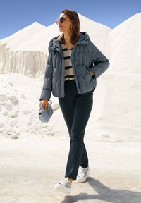 Gequiltede grijze pufferjack met capuchon, gestreepte beige en zwarte top, donkere broek en gestreepte sneakers; met een lichtblauwe handtas.