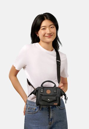 Young woman wearing white t-shirt and jeans, carrying black crossbody bag with front pocket and leather accents, smiling gently.