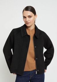 Čierna stredne dlhá bunda s gombíkovým zapínaním, zipsom a golierom. Mäkká textúra s mierne oversized strihom, doplnená hnedým topom.