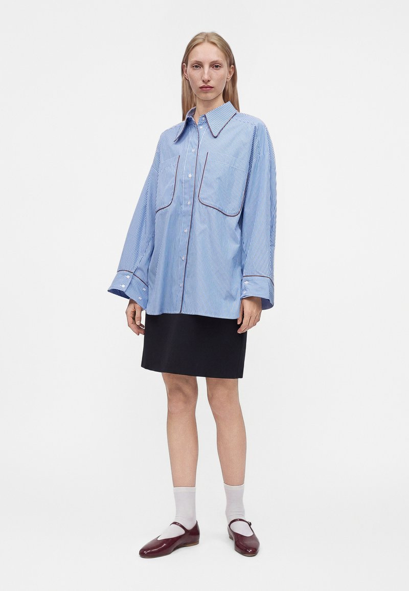 Femme portant une chemise boutonnée oversize à rayures bleues, une jupe noire, des chaussettes blanches et des chaussures Mary Jane rouge foncé, debout sur fond blanc.