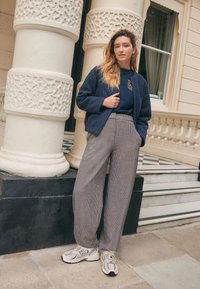 Navy textured jacket over a dark tee with logo, paired with wide-check, tailored trousers and beige sneakers featuring a logo accent.