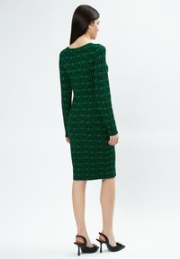 Grünes Strickkleid mit geometrischem schwarzen Muster, langen Ärmeln und knielanger Schnittführung, kombiniert mit schwarzen High Heels. Glatte Textur.