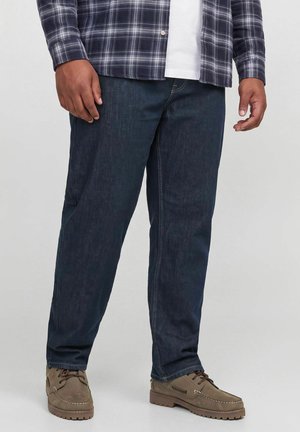 Homme portant un jean bleu foncé, des chaussures marron à lacets, une chemise blanche et une chemise à manches longues à carreaux, debout devant un fond uni.