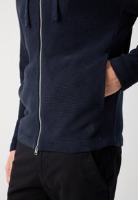 Felpa con zip di colore blu navy in tessuto testurizzato, con tasche frontali e logo discreto. Presenta una chiusura con zip e polsini aderenti. Abbinata a pantaloni neri.