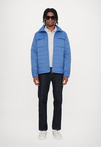 Light blue puffer jacket with a high collar, two front pockets, layered over a gray polo, paired with dark jeans and white sneakers.