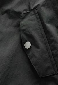 Tessuto nero con una texture liscia, caratterizzato da una patta per la tasca e un bottone a pressione in metallo argentato. Linee pulite e design minimale.