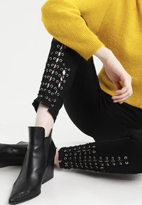 Černé strečové kalhoty s detaily šněrování na dolních nohavicích, spojené s špičatými černými kotníkovými botami a žlutým pleteným svetr.