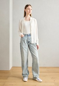 White, lightweight button-up shirt over a white top, paired with loose, light blue denim jeans and multi-colored sneakers. Wood floor background.
