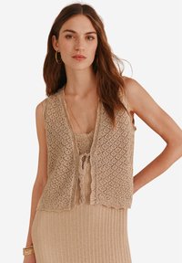 Gilet en crochet beige avec un bord festonné, un motif texturé et une ouverture sur le devant. Porté par-dessus une robe à côtes assortie, avec un décolleté rond.