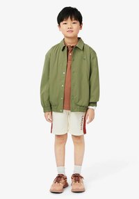 Giacca leggera verde oliva, polo marrone, pantaloni corti crema con dettagli rossi e sneakers marroni. Design semplice, vestibilità casual e polsini elasticizzati.