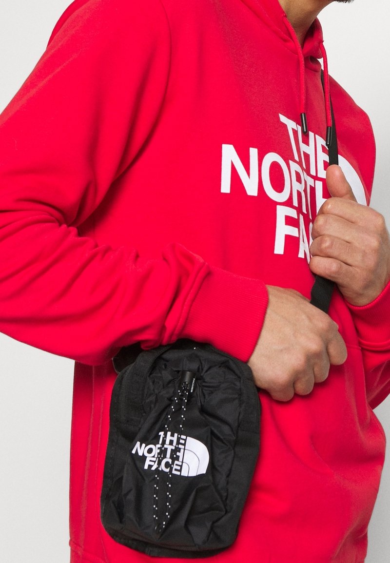 Borsa leggera con coulisse di colore nero, con il logo "THE NORTH FACE" in bianco. Indossata sopra un maglione rosso con un grande logo bianco.