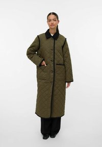 Kvinde iført en lang, quilted olivengrøn frakke med sort kant og knapper, stående foran en ensfarvet hvid baggrund.