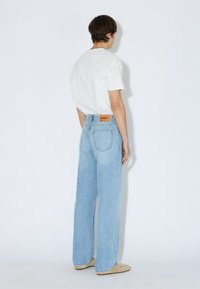 Light blue denim jeans with a wide-leg design and a faded finish. Features a leather back patch and two rear pockets. Worn with a white T-shirt.
