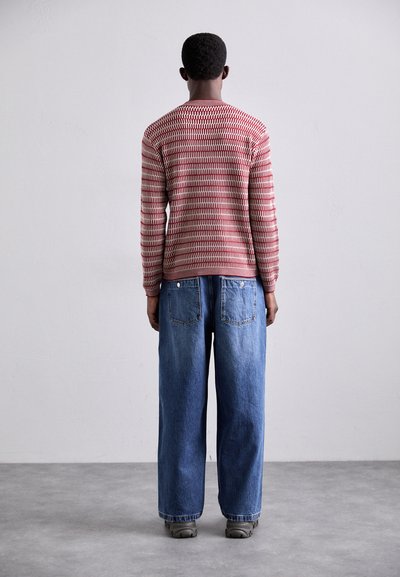 Rood en roomkleurig gebloemd longsleeve sweater gecombineerd met losse blauwe jeans. Model staat met de rug zichtbaar tegen een lichte, neutrale achtergrond.