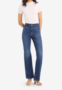 Mujer con top de manga corta rosa claro, vaqueros azules de talle alto con costuras diagonales y sandalias blancas de tacón con tiras, de pie con las manos detrás de la espalda.