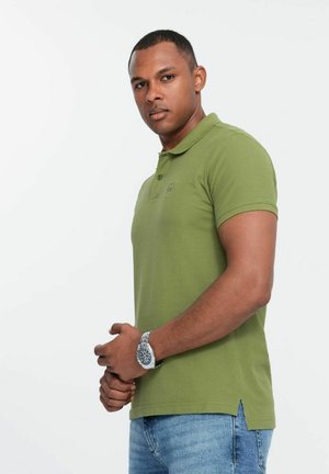 Camisa polo verde oliva de manga corta, con dos botones en el cuello y un pequeño logo en el pecho. Llevada con jeans de color azul claro.