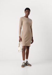 Beige stickad klänning med kabelmönster, långa ärmar, ribbad krage och nederkant. Matchad med grå sneakers och en vit väska.