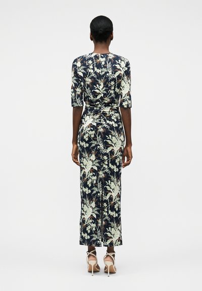 Γυναίκα με σκουρόχρωμο δέρμα που φορά φόρεμα midi σε στενή γραμμή με φλοράλ σχέδιο σε μπλε ναυτικό χρώμα και μπεζ ψηλοτάκουνα σανδάλια, όρθια με την πλάτη προς τον θεατή.