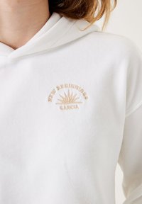 Sudadera blanca de tela suave, con el texto bordado en beige "Nuevos Comienzos" y un diseño de sol encima de "Garcia" en el pecho.