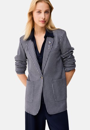 Giovane donna con capelli biondi che indossa una camicia blu navy e un blazer grigio a righe sottili con maniche arrotolate e spille decorative sul rever.