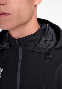 Chaqueta ligera negra con un alto cuello, cierre de mitad de cremallera y capucha ajustable. Tejido texturizado con un brillo sutil, que presenta un detalle de logo.