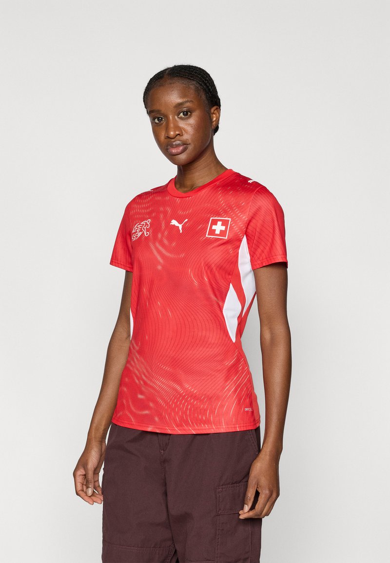 Κόκκινη αθλητική φανέλα με λευκές λεπτομέρειες, στρογγυλή λαιμόκοψη και δυναμικό κυματιστό μοτίβο. Φέρει τη σημαία της Ελβετίας και το λογότυπο της Puma στο στήθος. Κοντά μανίκια.