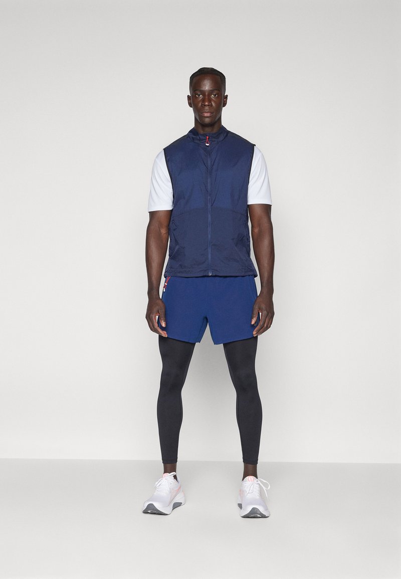 Tenue de sport pour homme comprenant un gilet navy, une chemise blanche à manches courtes, un short bleu, un pantalon de leggings noir et des chaussures blanches avec des accents.