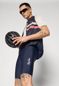 Los pantalones cortos de ciclismo azul marino tienen un asiento acolchado, combinados con una camiseta sin mangas azul con rayas rojas y blancas. Se sostiene un casco de ciclismo negro.