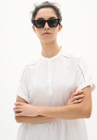 Bílá lněná košile s krátkým rukávem, s límečkem, a krajkovými detaily na ramenou, nošena s úhlovými černými slunečními brýlemi.