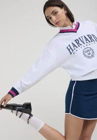 Felpa bianca corta con accenti blu e rosa, con logo "Harvard University". Gonna navy con piping laterale e scarpe nere con cinturini.