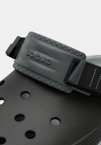 Zbliżenie na czarny but Crocs z teksturowanym, regulowanym paskiem z logo Crocs oraz otworami wentylacyjnymi z przodu.