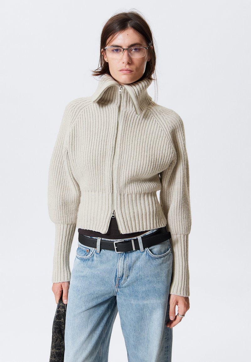Beige Rippstrickjacke mit hohem Kragen, durchgehendem Reißverschluss und cropped Design. Kombiniert mit Jeans und einem schwarzen Gürtel, der in einer Hand gehalten wird.